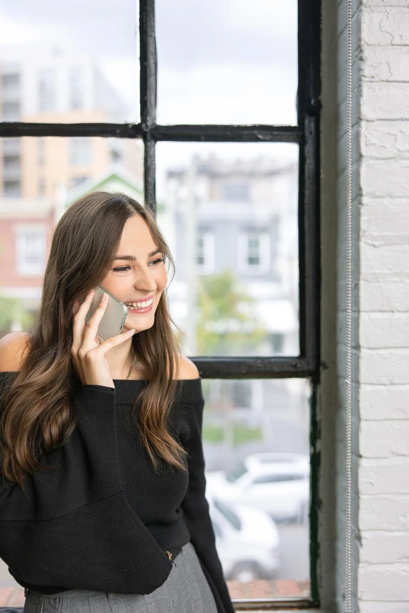 A young woman with long brown hair smiling and talking on her cell phone near a large window with black framing, with a cityscape of buildings and parked cars outside.