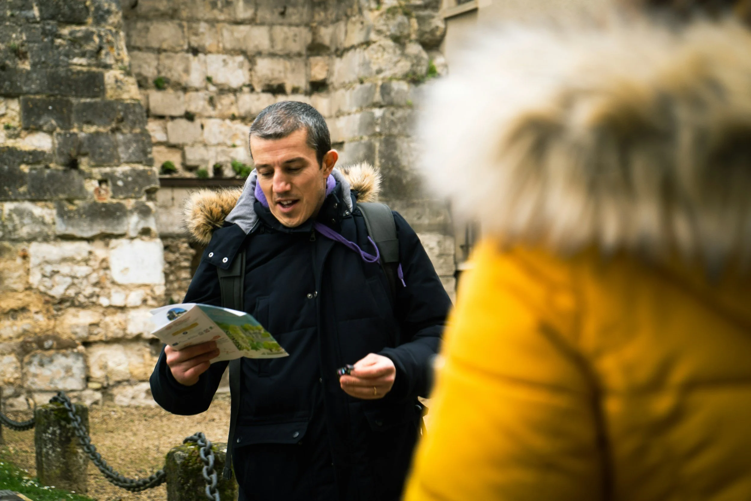 A man wearing a black jacket with fur trim, holding a map and looking at it, with another person in a yellow jacket in the foreground. They are outdoors near a stone wall.
