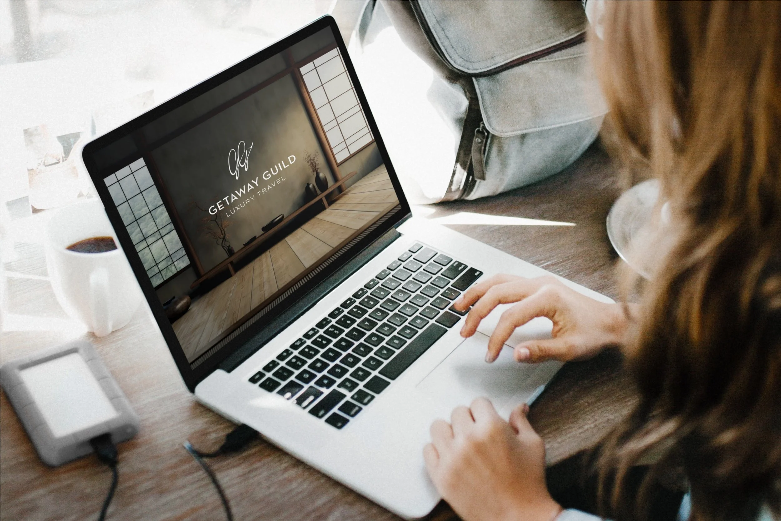 A person using a laptop on a wooden table, with a coffee mug and a portable charger nearby, and a backpack on the side. The laptop screen displays 'Getaway Guild Luxury Travel'.