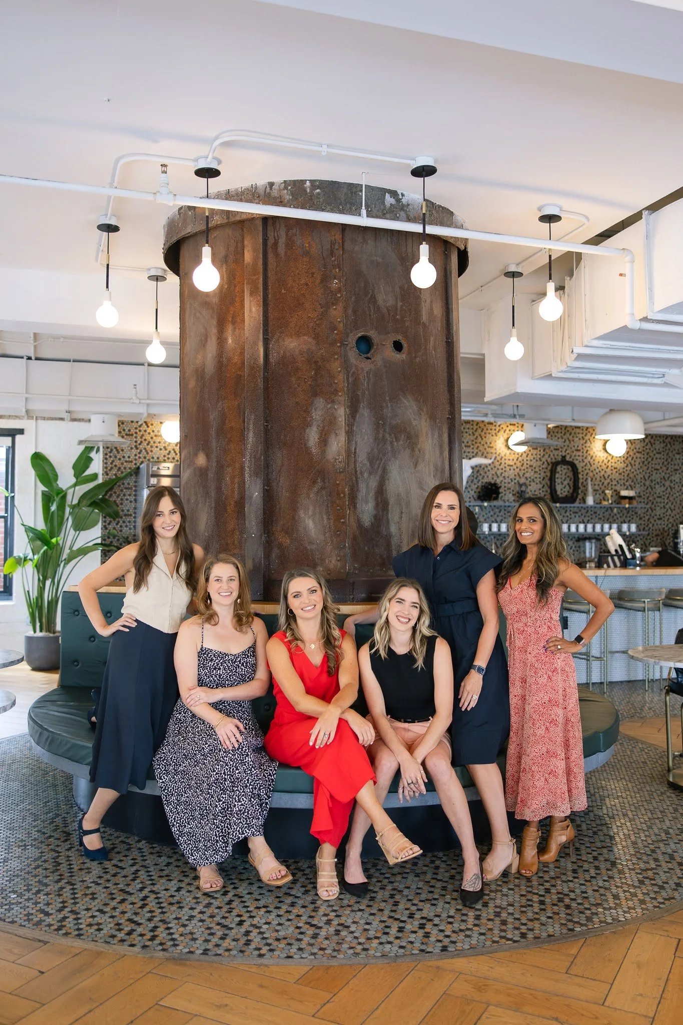 Group of seven women smiling and posing together in a stylish indoor space with modern decor, a large rusty industrial pipe feature, and bright lighting.