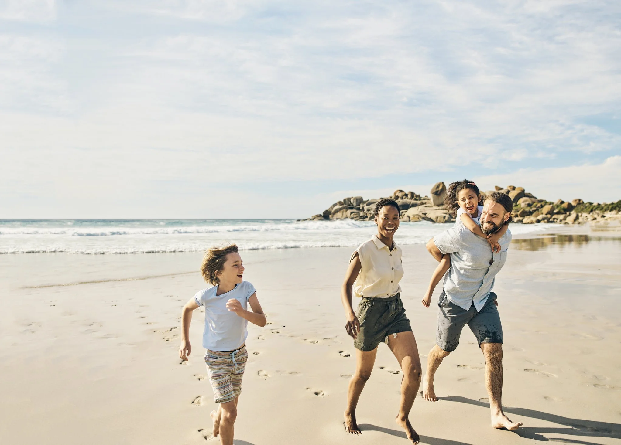 Family running and laughing on the beach, with rocks and ocean in the background.