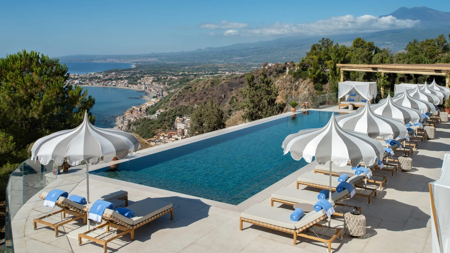 Infinity pool with white umbrellas and lounge chairs overlooking a coastal town and distant mountains.