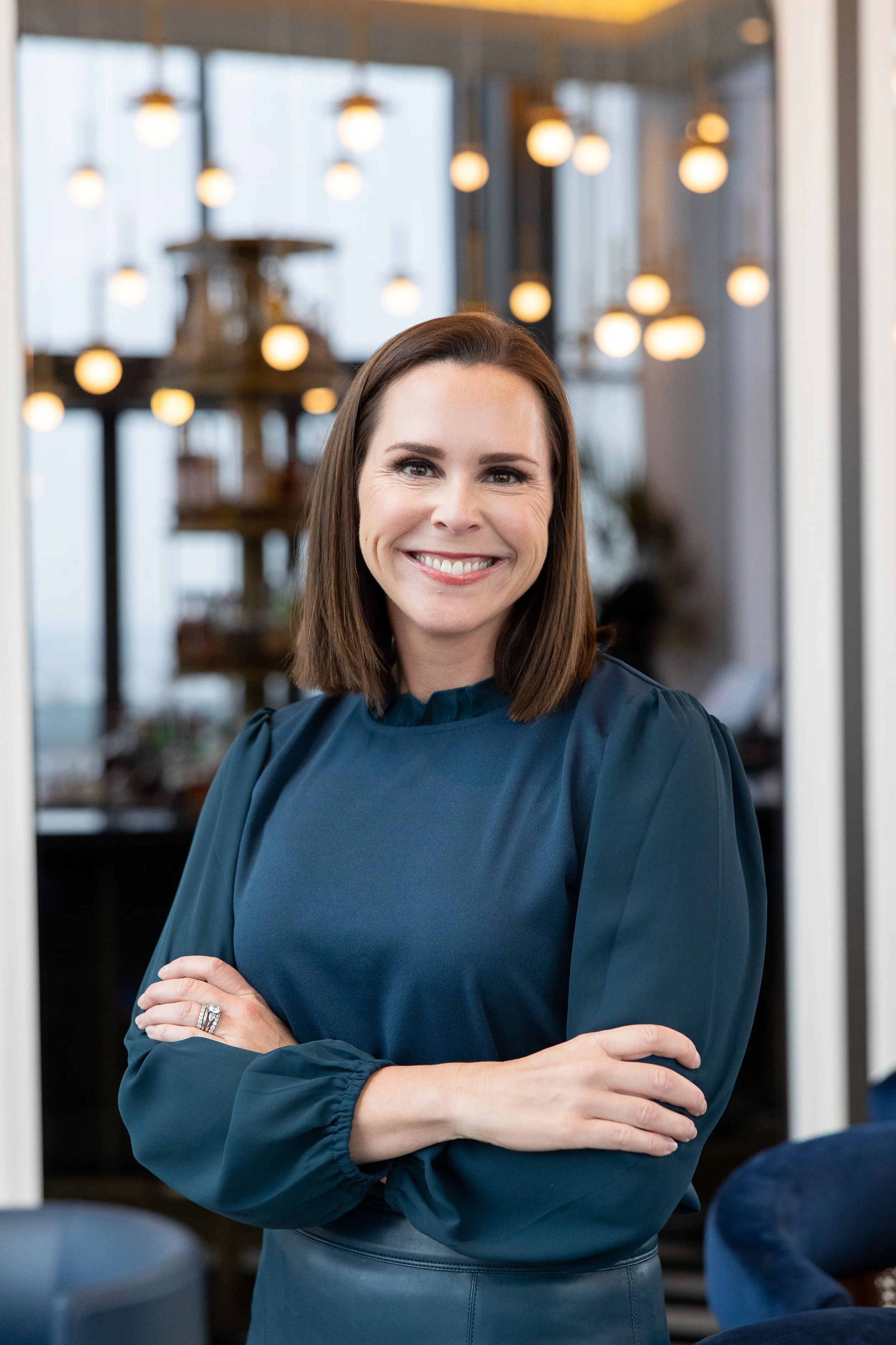 A smiling woman with brown hair in a blue blouse standing with crossed arms in a modern, well-lit indoor space with hanging globe lights and a decorative shelving unit in the background.