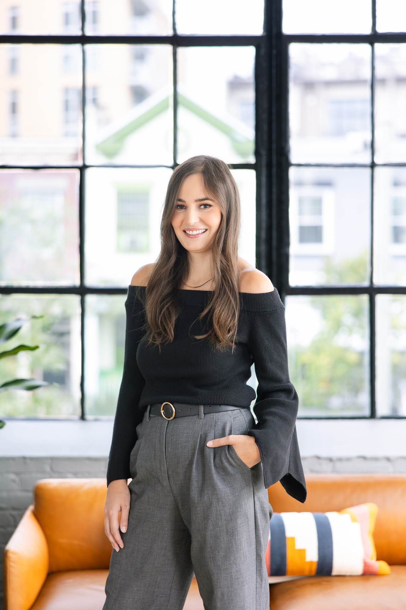 Young woman with long brown hair smiling, wearing a black off-shoulder sweater and gray high-waisted pants with a black belt, standing in front of large industrial-style window in a modern, brightly lit interior.