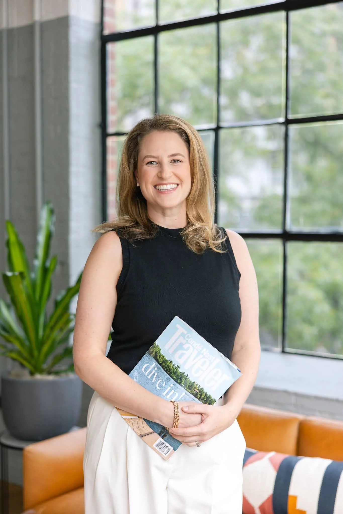 A woman with shoulder-length light brown hair wearing a black sleeveless top and white pants, smiling and holding a magazine called 'Travel' with the title visible, standing in a modern, brightly lit room with large windows and green plants.