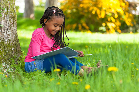 Young girl sitting on grass next to a tree, reading a book outdoors in a park during fall.