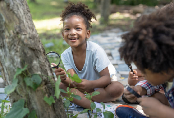 Two children outdoors examining a tree with magnifying glasses, one girl smiling and sitting near the tree, a boy looking closely at the tree with a magnifying glass.