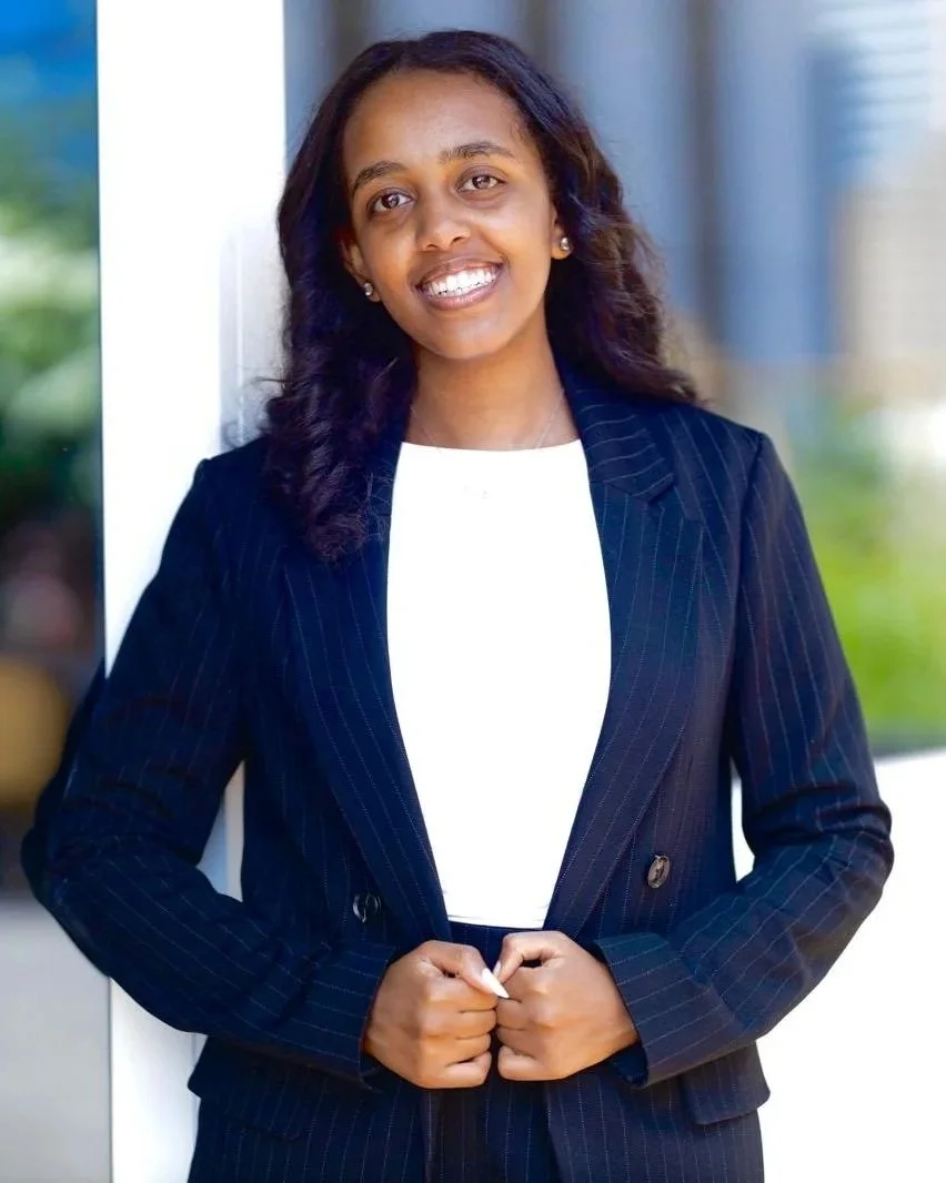 A woman with dark, wavy hair, wearing a navy pinstripe blazer over a white top, standing indoors and smiling at the camera.