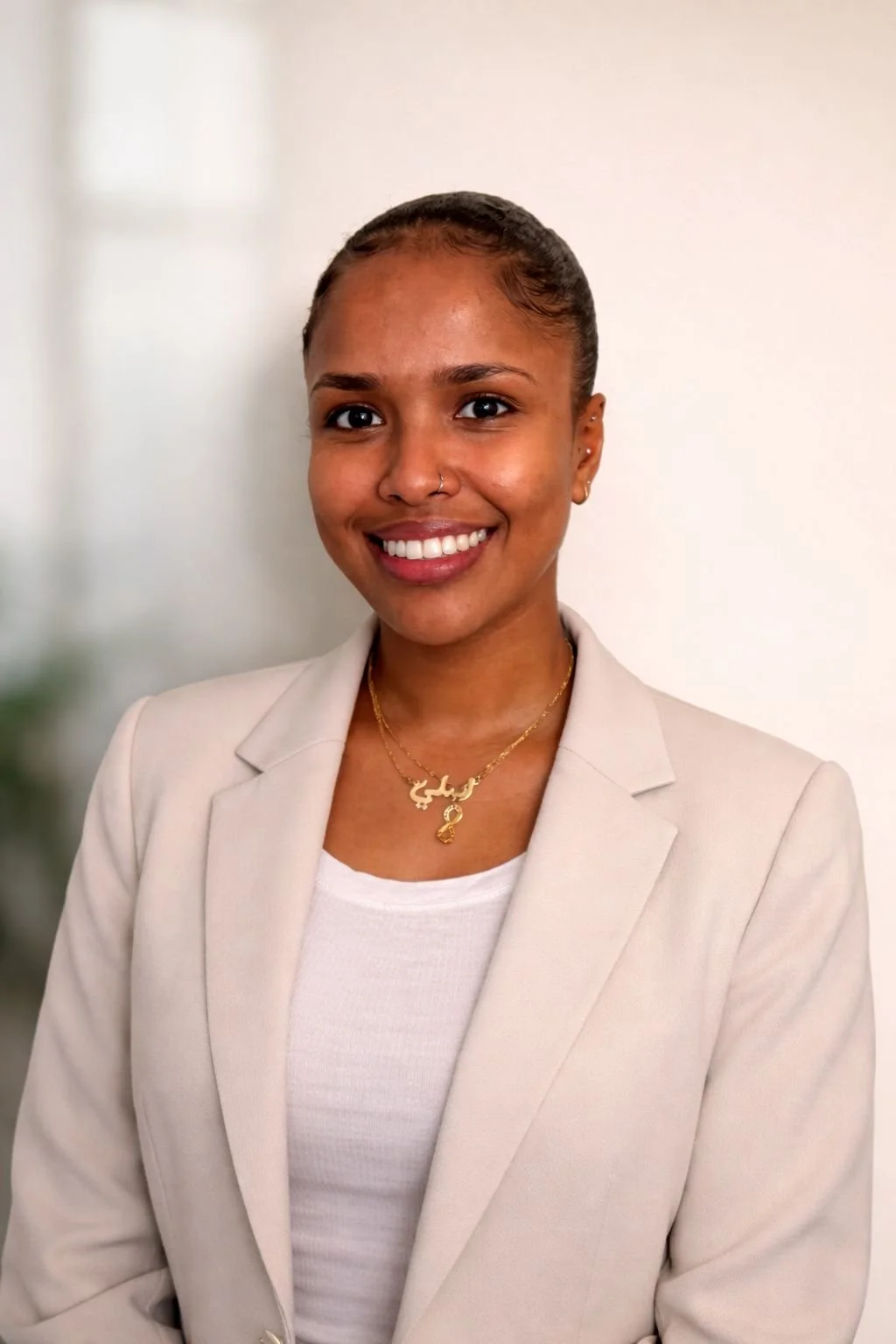A young woman with a bright smile, wearing a beige blazer, white top, and gold jewelry, standing against a plain background.