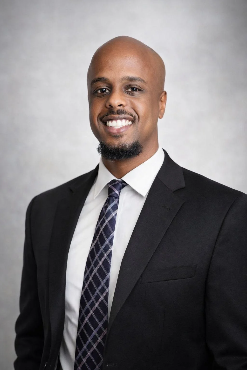 Professional portrait of a smiling man wearing a black suit, white shirt, and patterned tie, against a plain background.