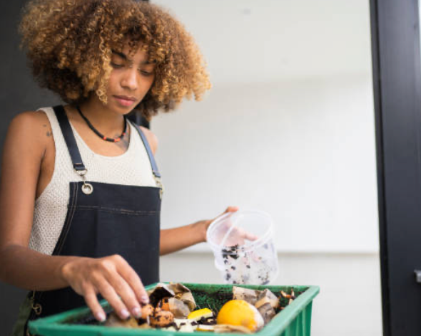 Young woman with curly hair and denim overalls sorting trash into a green recycling bin