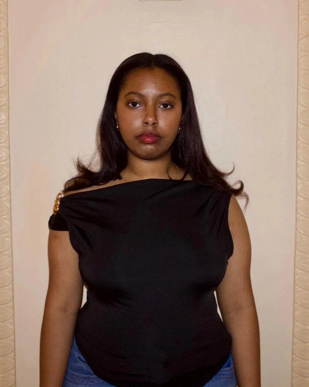 A young woman with dark hair and a serious expression, wearing a black off-the-shoulder top and blue jeans, standing against a plain background.
