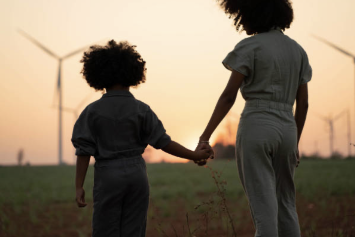 Two girls holding hands in a field during sunset, with wind turbines in the background.