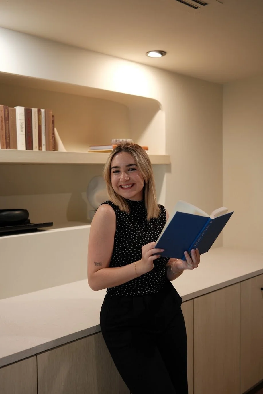 A young woman with shoulder-length blonde hair smiling and holding an open book, standing in a room with a beige bookshelf and countertop.