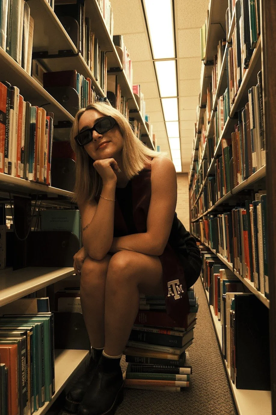A young woman wearing black sunglasses, a sleeveless dark top, and a maroon scarf with a university logo, sitting on a stack of books in a library aisle. She is smiling slightly, with her chin resting on her hand, surrounded by bookshelves filled with books.
