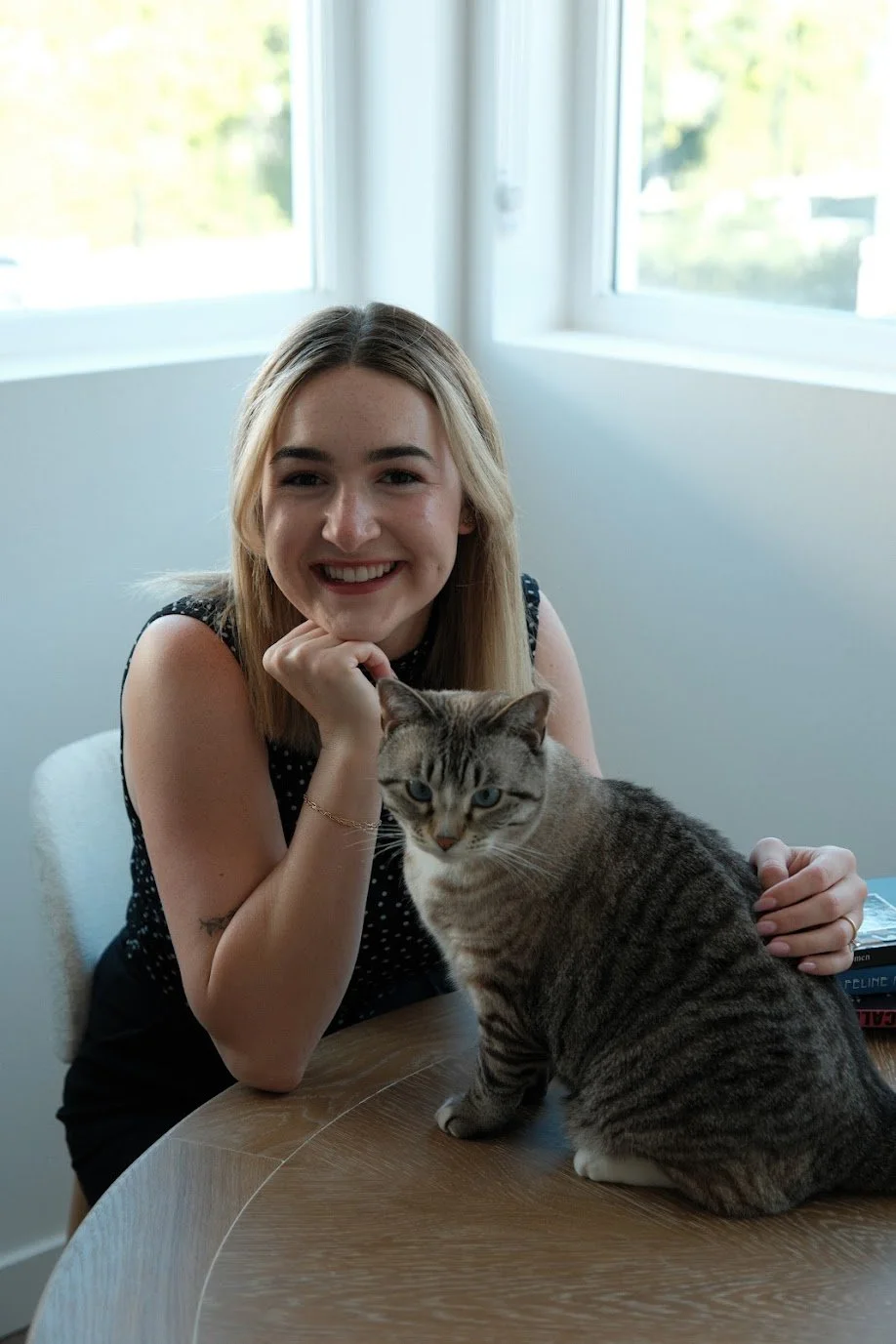 A young woman with shoulder-length blonde hair smiling while sitting at a wooden table with a tabby cat. The woman’s chin resting on her hand and the cat is sitting on the table in front of her. Bright natural light coming through windows behind them.