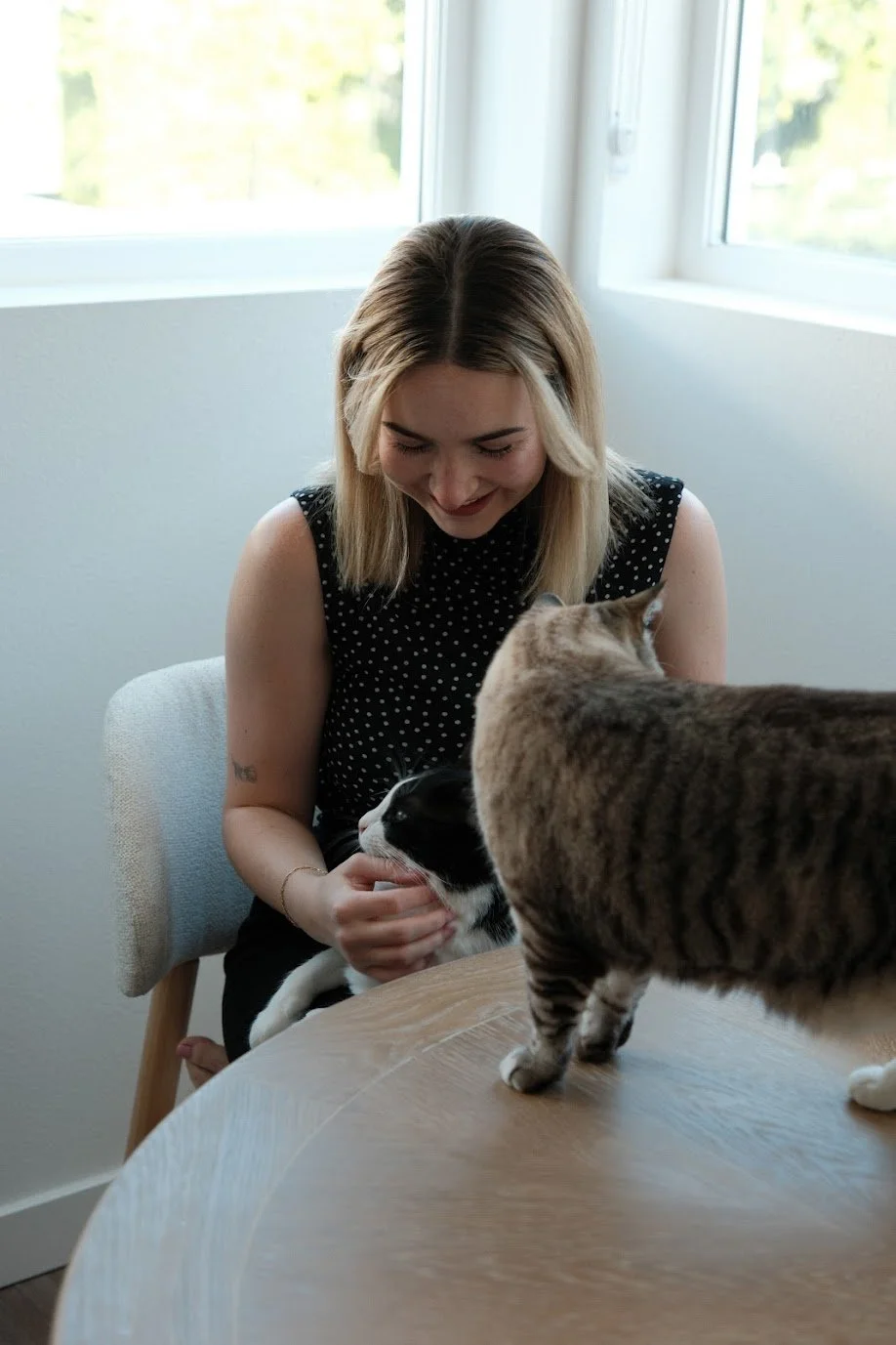 A woman with blonde hair and a black polka dot sleeveless top smiling while petting two cats, one black and white and dirty, and the other tabby, on a wooden table near a window.