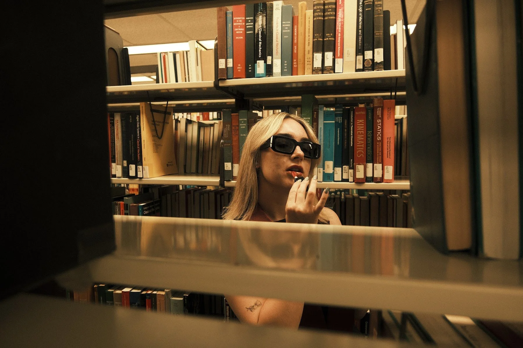 A young woman with blonde hair, wearing sunglasses, is in a library aisle between bookshelves. She is holding a cigarette near her mouth, and part of her arm with a tattoo is visible.