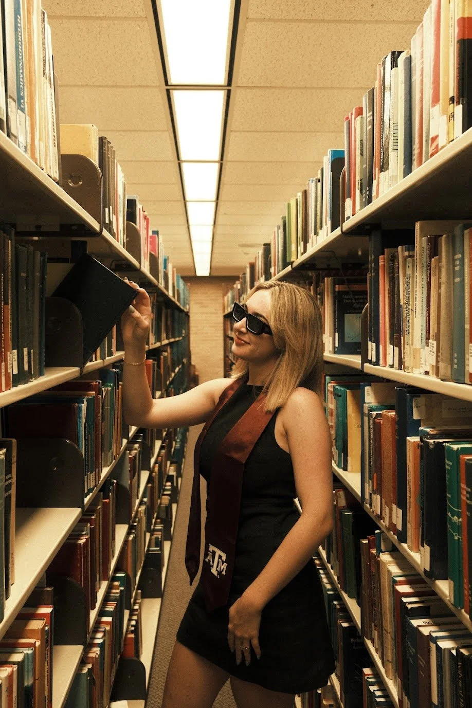 A woman with blonde hair wearing sunglasses, a black sleeveless dress, and an academic stole standing in a library aisle, reaching for a book on a shelf.