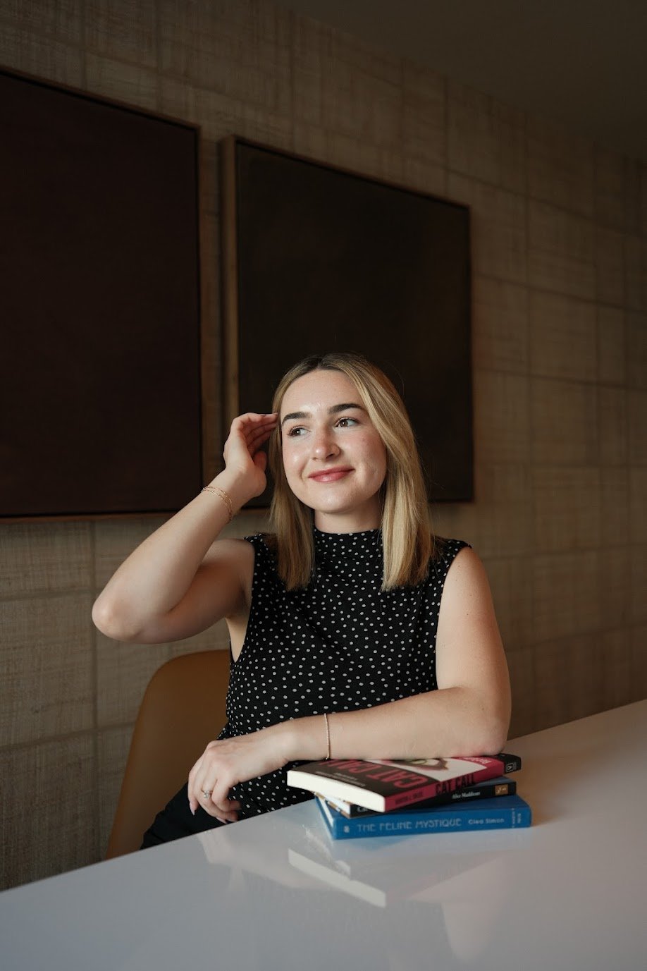 A young woman with blonde hair and fair skin, wearing a black sleeveless polka-dot top, is sitting at a white table. She has a small smile, and her left arm rests on the table while her right hand touches the side of her head. There are three books stacked on the table in front of her. The background features a wooden wall and two blank blackboards.