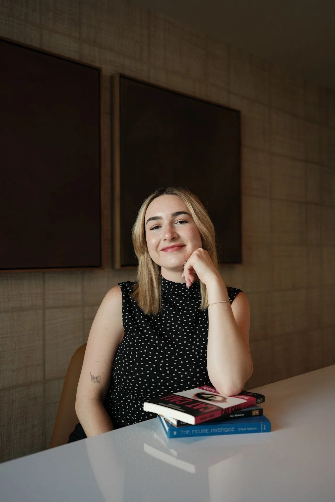 Young woman with blonde hair, wearing a black sleeveless polka-dot top, sitting at a white table with three books in front of her, smiling and resting her chin on her hand.