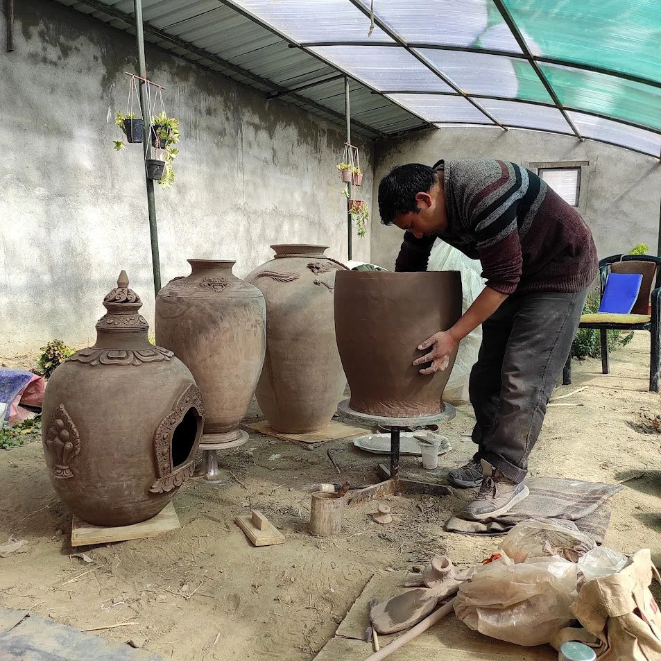 A man shaping a large clay pot on a pottery wheel, surrounded by several finished and unfinished clay pots in a greenhouse or workshop setting.