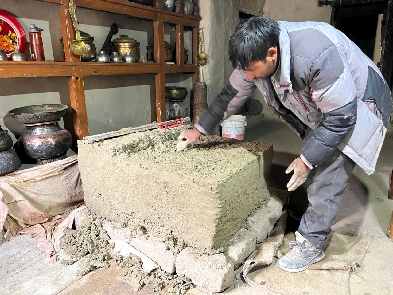 A man working on a large stone block, smoothing its surface with a trowel in a workshop with shelves holding various metal pots and tools.