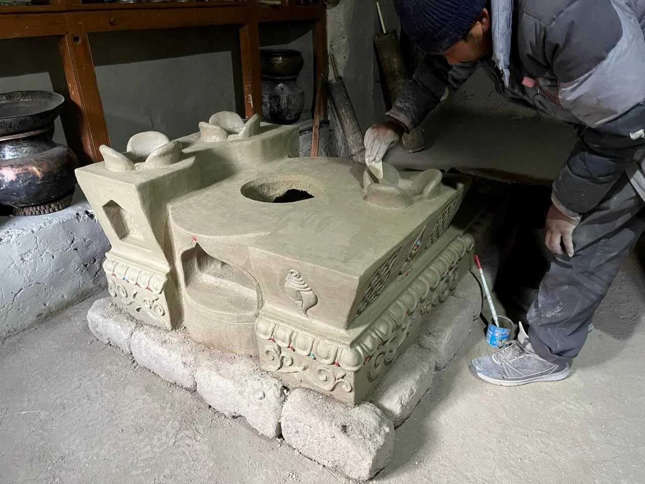 A person working on a carved clay stove (thap), with decorative designs and a large central hole, in a workshop setting.