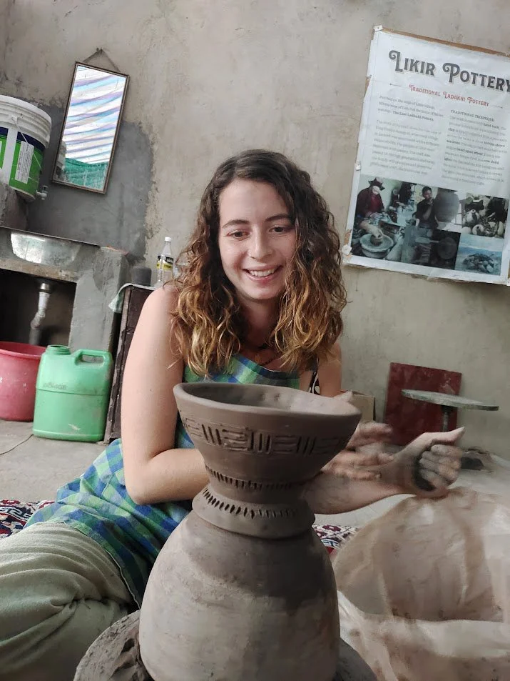 A young woman with curly hair smiling while working on a pottery sculpture in a workshop.