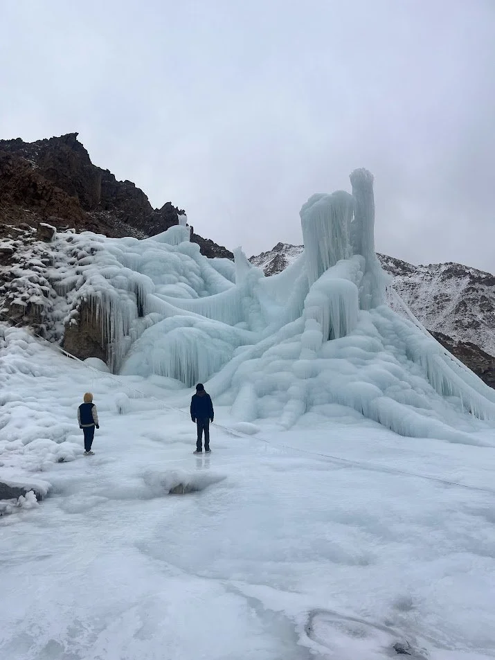 Two people stand on a frozen landscape in front of an icy artificial glacier formation with icicles and ice layers, set against a mountain and cloudy sky.