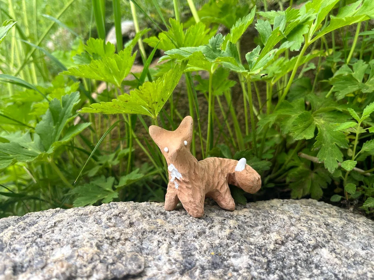 A small brown and beige fox figurine with white paint chipping on its body, placed on a textured gray rock amidst green leafy plants.