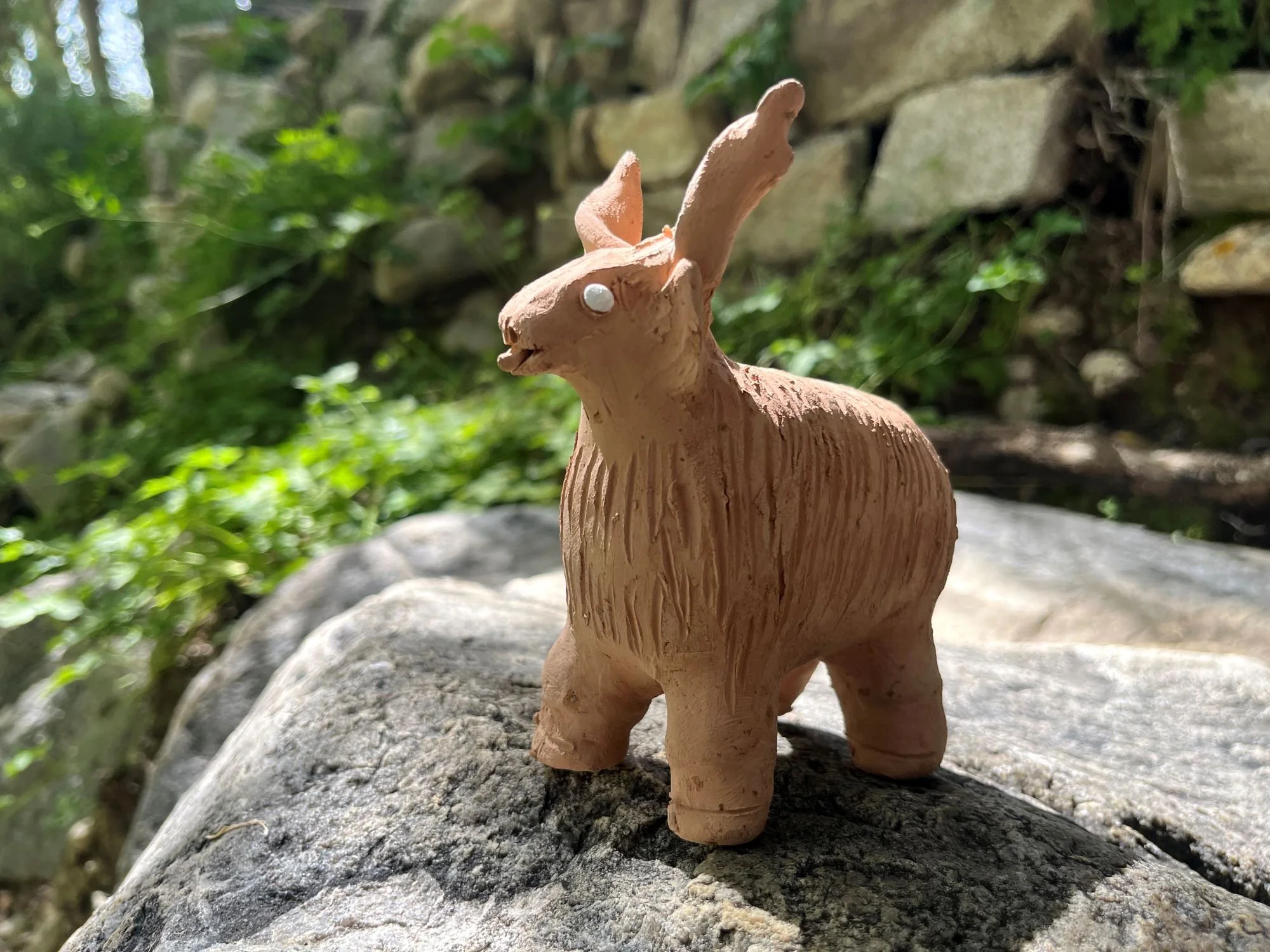 A small, handcrafted clay sculpture of a yak with long ears, standing on a rock outdoors with green foliage and a stone wall in the background.