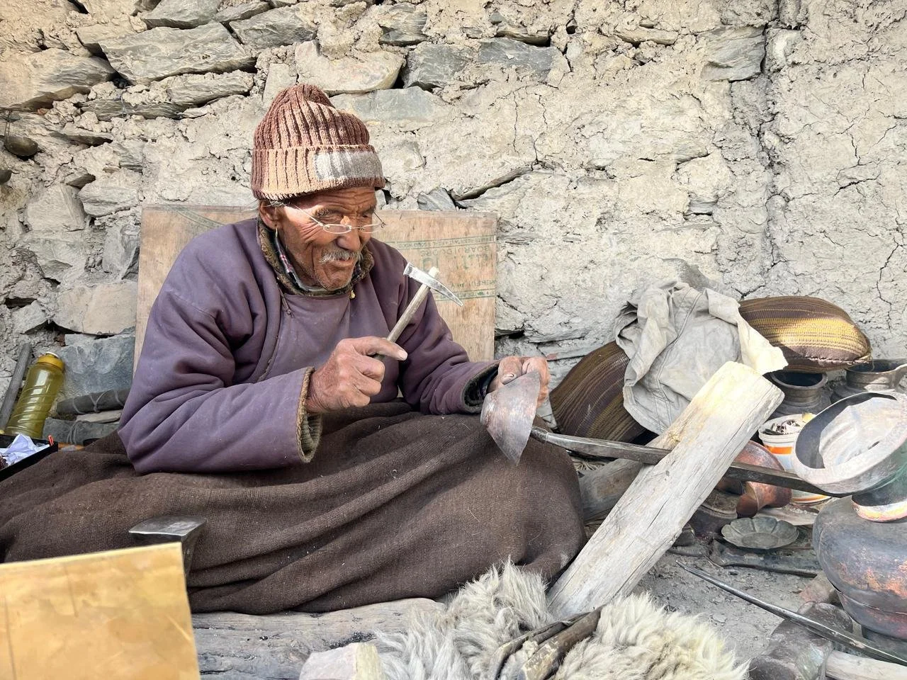 An elderly man with glasses and a beanie hat sits on the ground, working as a blacksmith using a hammer and chisel. The scene is outdoors against a stone wall, with various tools, pottery, and supplies scattered around him.