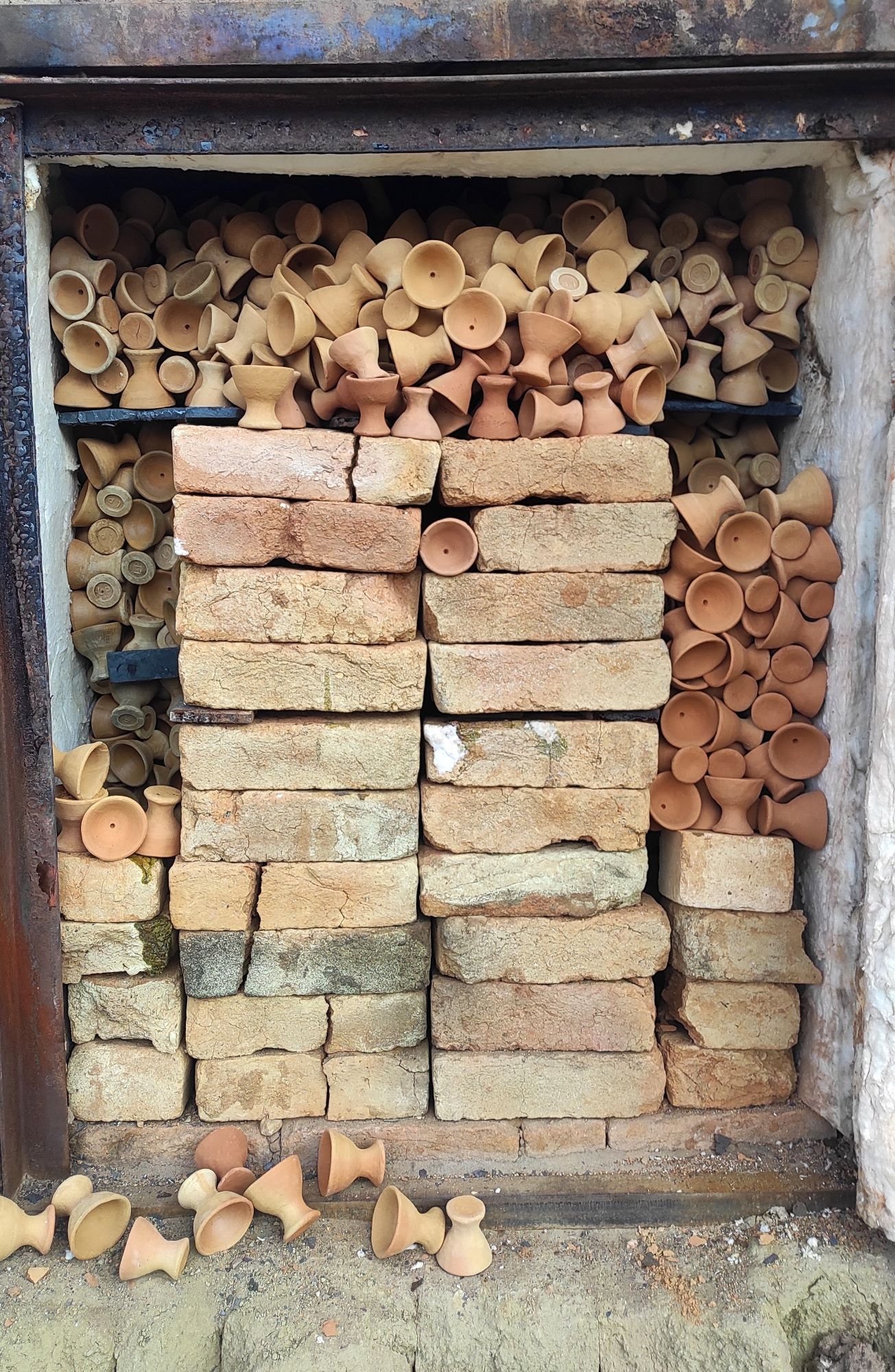 A collection of unglazed clay pottery vessels stored in a brick and metal frame. Some scattered pottery pieces are on the ground in front of the storage area.