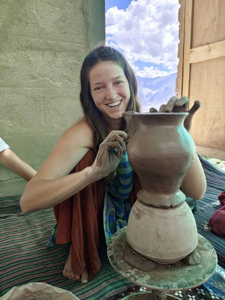 A woman with long dark hair smiling, holding a clay pot she's shaping on a pottery wheel, with a mountain landscape and blue sky in the background.