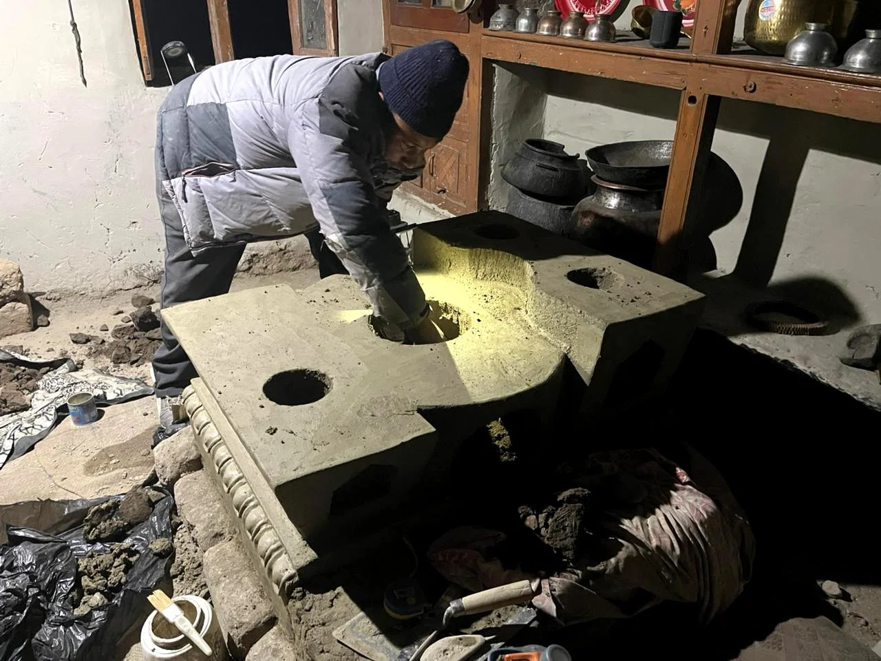 A person working on a s clay stove, wearing a winter jacket and a beanie, with tools and construction materials around.
