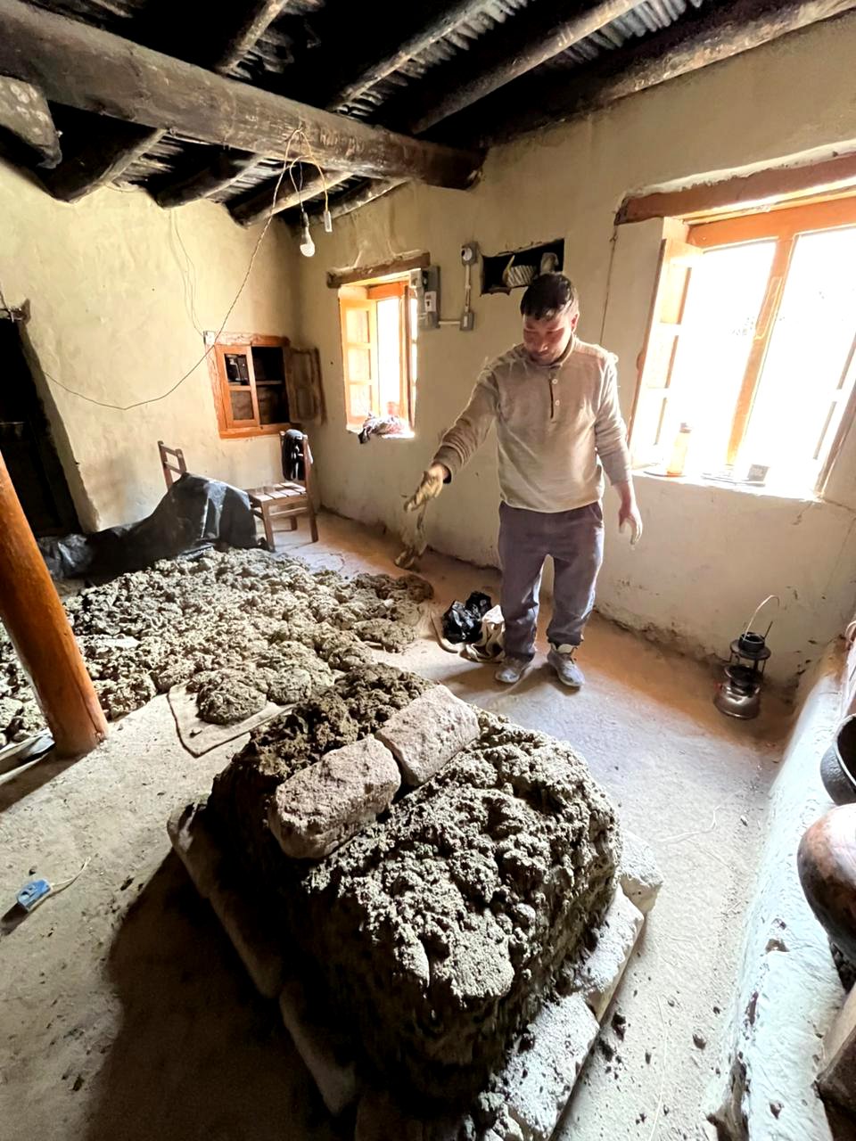 A man working with old mud bricks and mortar - making a earthern stove inside a rustic room with wooden beams, small windows, and minimal furnishings.