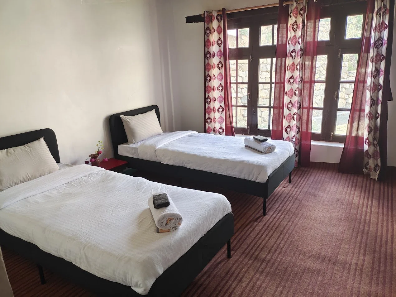 A hotel room with two single beds, each with white linens, pillows, and towel arrangements, situated near large window with red patterned curtains, and a small bedside table between the beds.