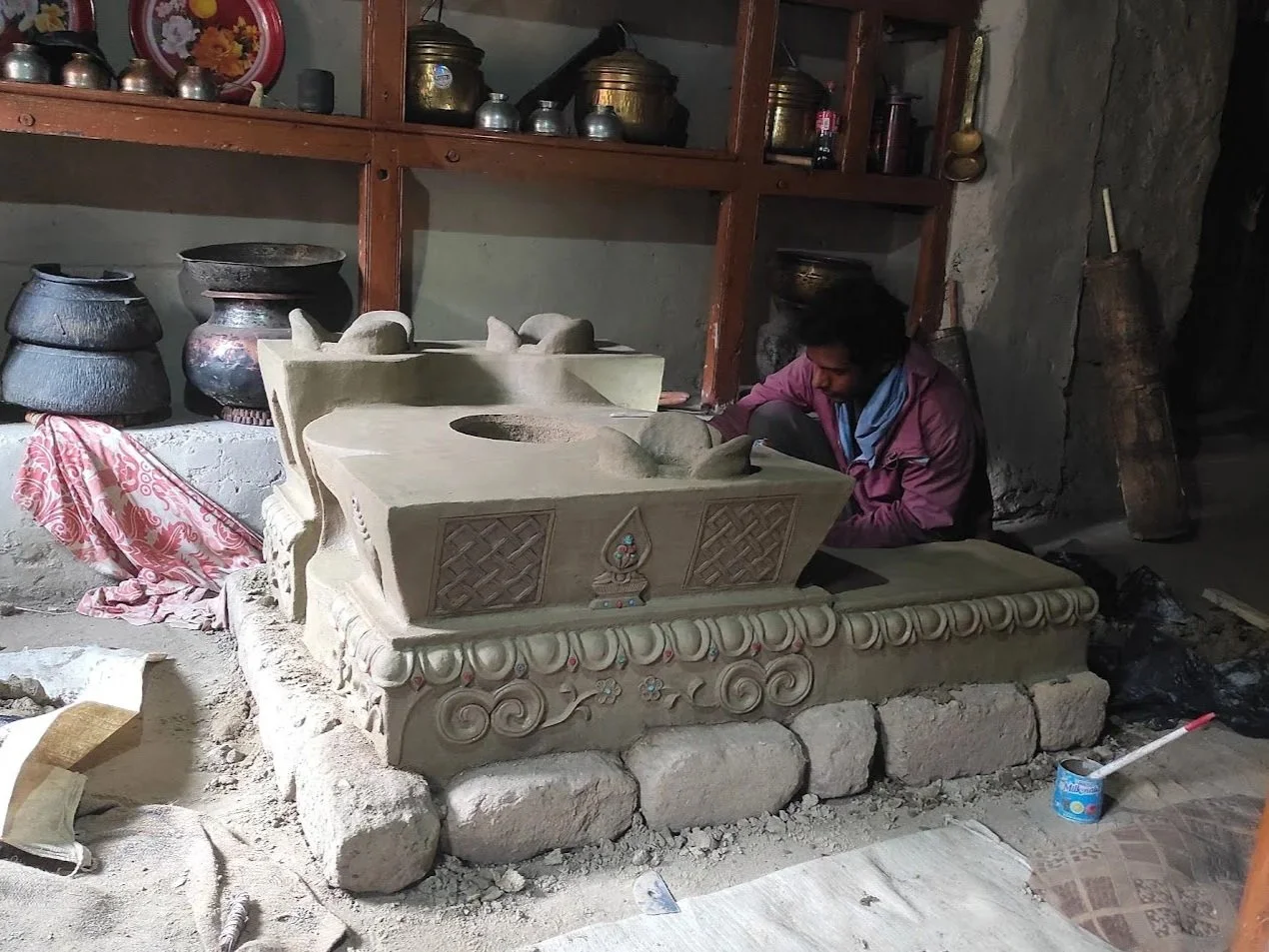 A person working on carving a traditional clay stove (thap). The sculpture features intricate designs and is placed on a stone base. Shelves in the background hold metal and ceramic pots.