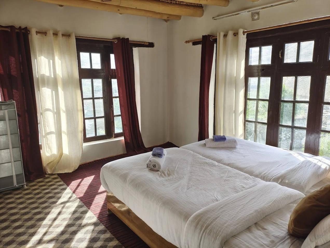 A bedroom with two beds, each with white linens and rolled towels, large windows with dark wooden frames, and light curtains, sunlight streaming in.