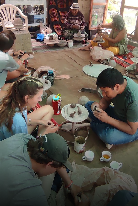 People sitting on the floor in a room, shaping clay pottery, with pottery tools, cups, and supplies scattered around.