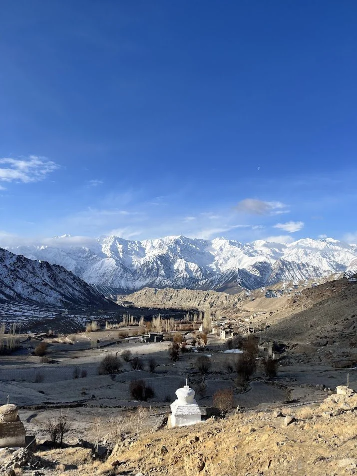 Snow-covered mountains under a clear blue sky with a small village in the valley, and a white stupa in the foreground.