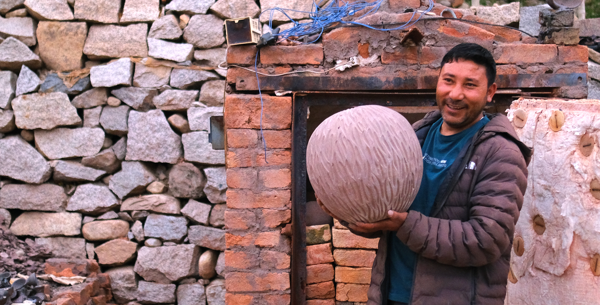 A man smiling and holding a large, round, textured clay object. He stands in front of a brick and stone wall, wearing a blue shirt and a brown jacket.