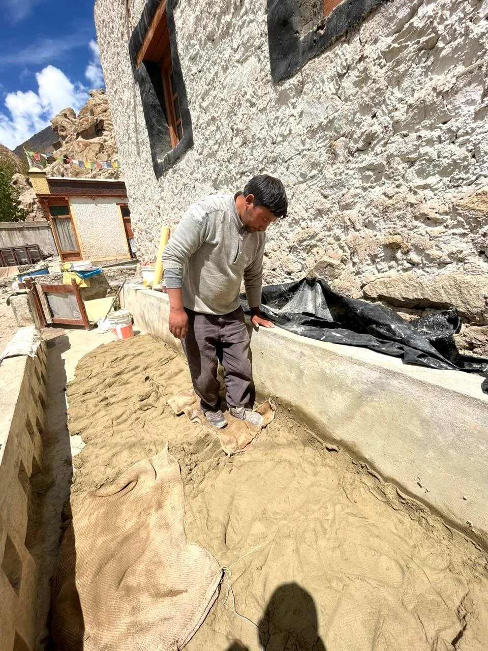 A man standing in a construction area with sand, next to a stone wall and a building. The sky is blue with some clouds.