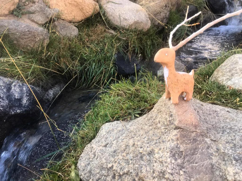Small decorative figurine of an antelope with white antlers, placed on a large rock near a stream, surrounded by grass and rocks.