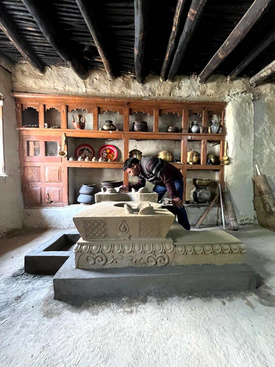 A man inspecting or working on a carved clay stove (thap) inside a rustic room with wooden shelves holding pottery and decorative items, wooden ceiling beams overhead, and a window on the left.