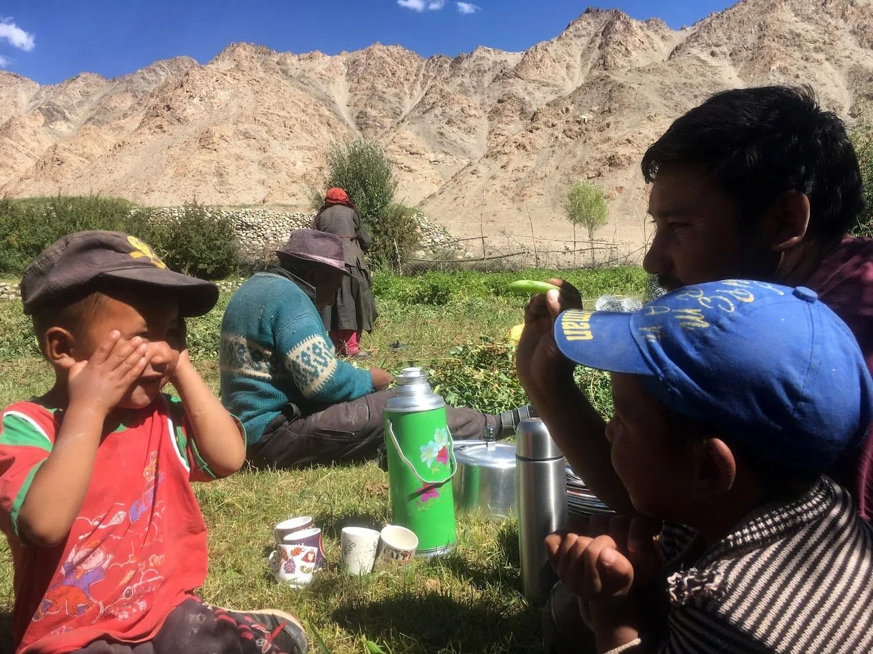 Children and a man sitting on grass in a mountainous area, having tea and snacks, with a woman in the background and dry mountains under a clear sky.