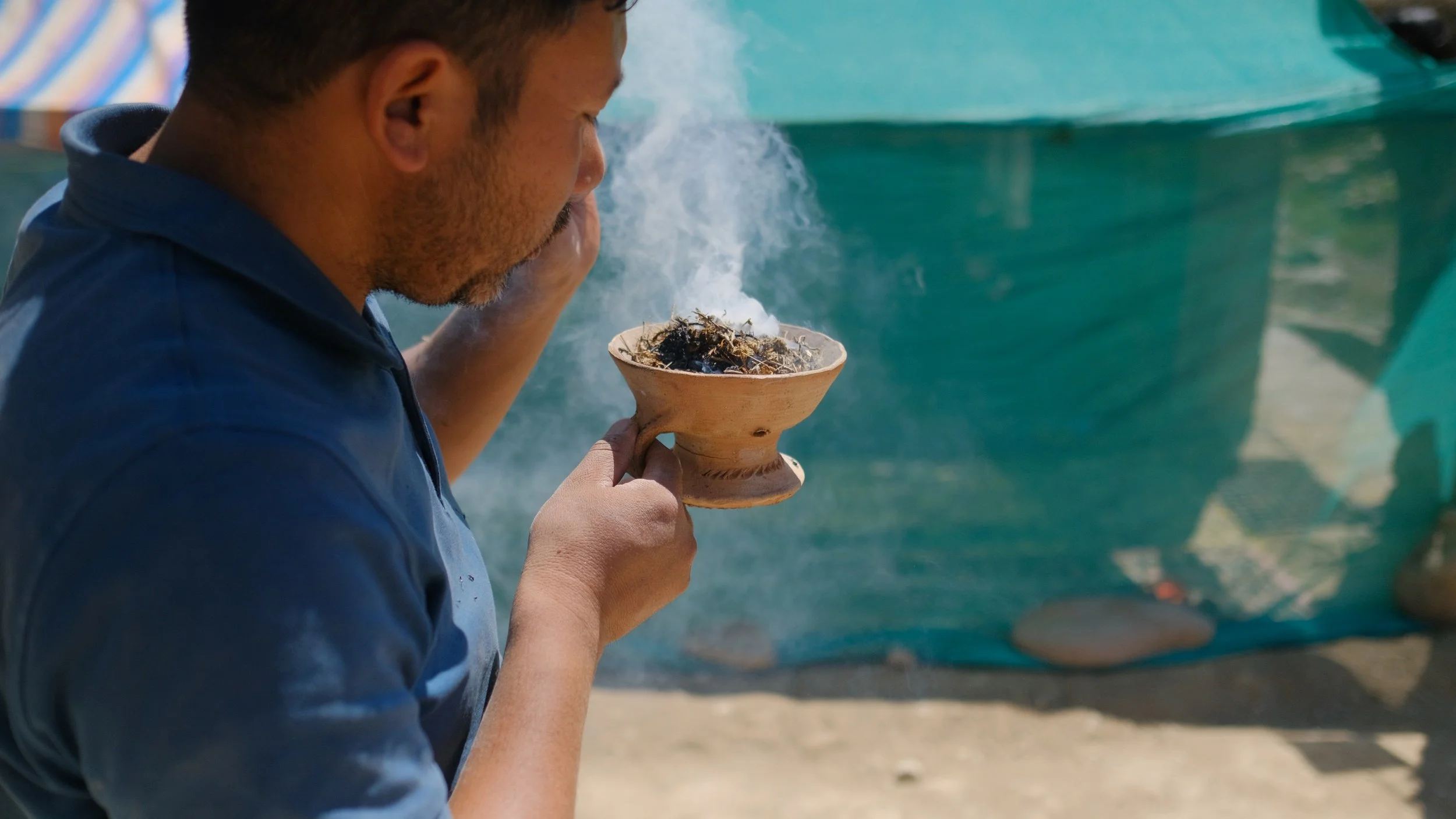 Rigzen namgail in a blue shirt blowing sr incense from a traditional pot with a rounded base and a narrow opening, with smoke rising from the pipe, outdoors near a green tarp or wall.