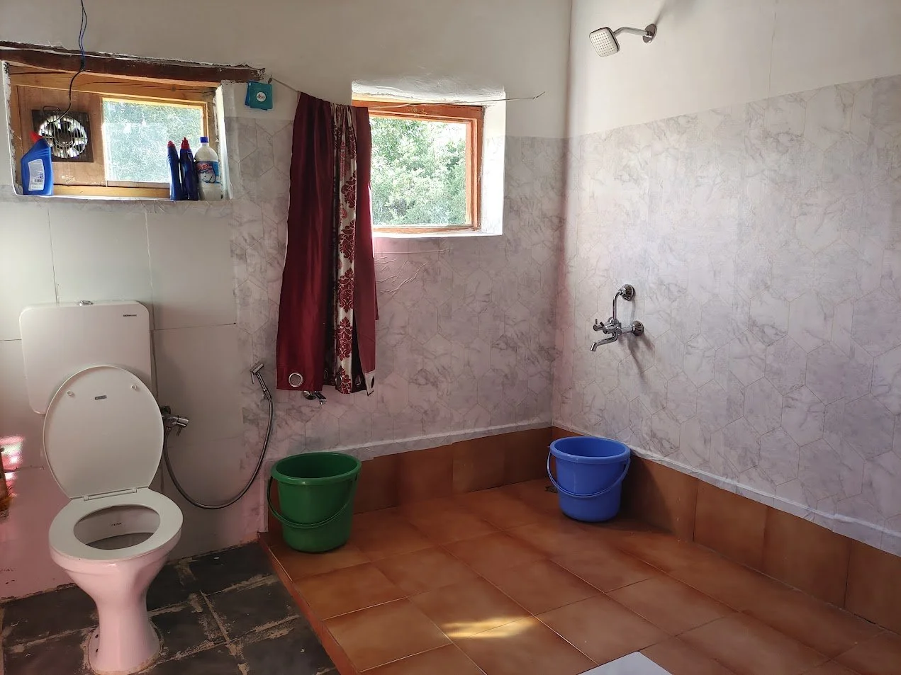 A simple bathroom with a toilet, two windows, a shower head, and two buckets on the floor. One window has a red and floral curtain, the other is uncovered. Bottles of cleaning supplies are on a small shelf by the window. The room has tiled walls and 