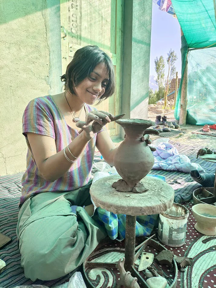 A woman working on a pottery project, shaping a clay vessel in a bright, open workshop with a makeshift outdoor setting.